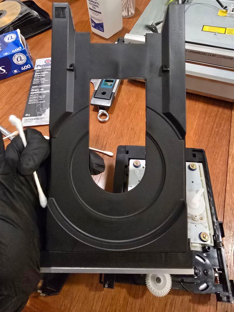 Disassembled Sony CD loading tray and cam gears on a workbench beside isopropyl alcohol, cotton swabs and synthetic grease